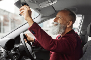 Elderly businessman in stylish suit getting ready before travelling by his auto, setting rearview...
