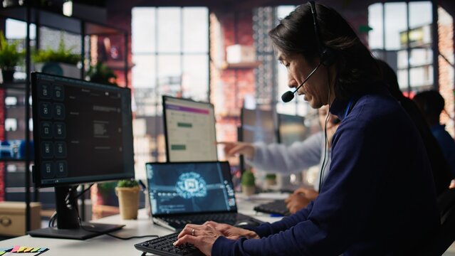Call center worker arriving at work, using artificial intelligence on laptop to enhance customer interactions. Asian senior agent entering customer service office, using AI technology, camera B