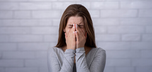Depressed millennial woman covering her face and crying against white brick wall. Sad young lady...