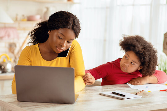 Bored Black Girl Demanding Attention From Her Busy Mom Working At Home, African American Freelancer Lady Using Laptop And Talking On Cellphone In Kitchen, Ignoring Her Lonely Child, Free Space
