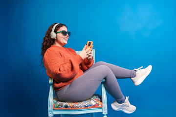 Plus size woman using mobile phone while listening music with headphones and wearing sunglasses, sitting on a chair