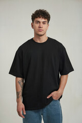 A studio portrait of a young man wearing an oversized black t-shirt and light blue jeans. The subject has short, tousled brown hair and visible tattoos on their left forearm. 