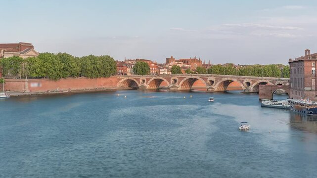 Garonne River and Pont Neuf timelapse with Port de la Daurade in downtown Toulouse, France. This Renaissance arch bridge reflects in the water under a blue sky with clouds. Waterfront with green trees