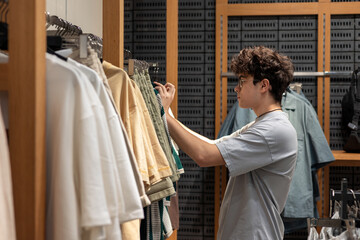 Teen boy choosing stylish t-shirt in clothing store. Young customer shopping trendy casual wear at fashion retail shop. Boy shopping alone in urban fashion shop. Copyspace 
