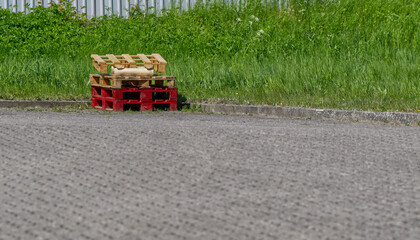 Red and pure wooden pallets are stacked on a driveway in front of a green area, the foreground and...