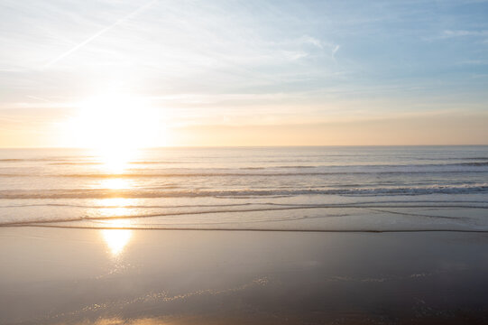 Sun is setting over calm ocean with soft waves reaching shore. Warm sunlight reflects on water creating bright glow. Sky is partly cloudy with soft blue and orange tones