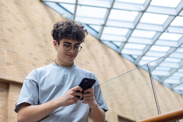 Smiling teenage boy using smartphone in modern urban space. Concept of youth lifestyle, digital...