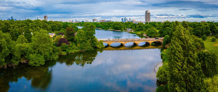 Aerial view of Hyde Park in London featuring the Serpentine lake, lush greenery, and a stone bridge with city skyline in the background