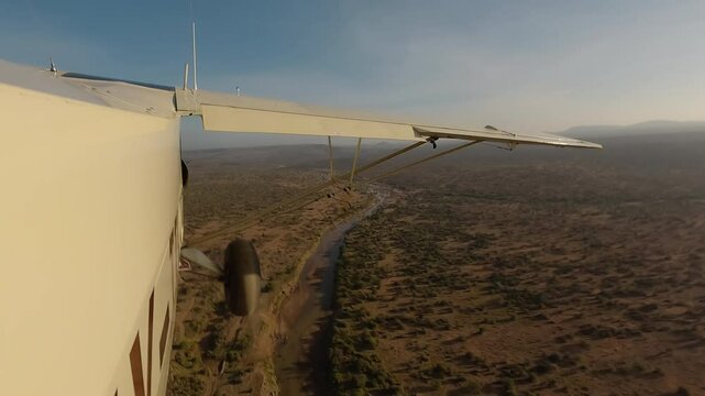 Aerial wide of small air plane side view flying over dry savannah with shrubs, river and road in a sunny morning in Kenya