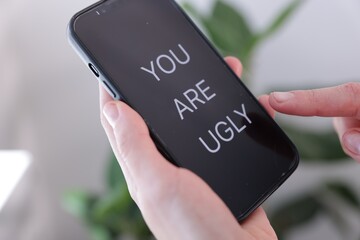 Cyber bullying. Woman holding smartphone with text You Are Ugly on blurred background, closeup