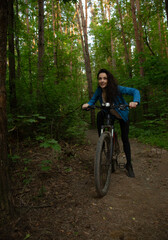 Fototapeta premium Beauty smiling woman biking on bicycle on spring trees green forest background, looking happy. Enjoying fun girl outdoors. Female cyclist riding in park in sport fashion clothes. Full lenght portrait