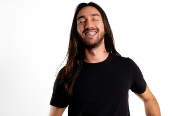 Latin man with long hair, close eyes, black t-shirt and braces laughs in front, on white background
