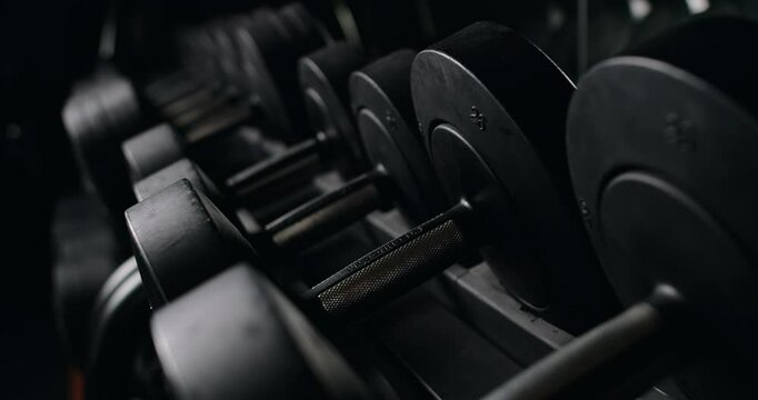 A low-light shot of a row of black dumbbells neatly arranged on a rack in a gym, focusing on the textures and contrasts shaped by ambient lighting. View of Weights on a Rack in a Dark Gym Setting
