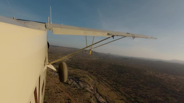 Aerial wide of small air plane side view flying over dry savannah with shrubs, river and road in a sunny morning in Kenya