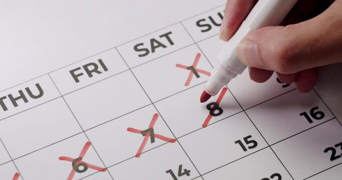 Close-up image of a person crossing off days on a calendar using a red marker pen, indicating the passage of time or counting down to an event. Person Crossing Off Days On Calendar With Red Marker Pen