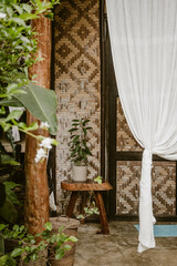Vertical image of a wooden stool with a tropical green plant outside on the porch of an aesthetic bamboo house made of natural materials at daytime on a holiday in the Philippines in Asia.