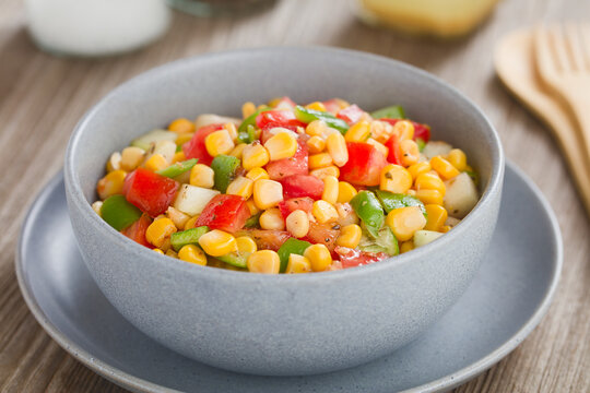 Colorful vegetable salad made of tomato, green pepper, cucumber and corn kernels, served in bowl, with vinaigrette in the back (Selective Focus, Focus in the middle of the image) 