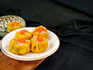 Yellow dimsum or Siomay chicken and shrimp variance with carrot above served on white plate,chopsticks, wooden cutting board, in black background
