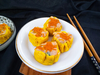 Yellow dimsum or Siomay chicken and shrimp variance with carrot above served on white plate,chopsticks, wooden cutting board, in black background
