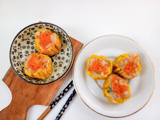 Yellow dimsum or Siomay chicken and shrimp yellow dimsum chicken and shrimp variance with yellow dimsum with carrot above served on white plate,chopsticks, wooden cutting board, in white background.
