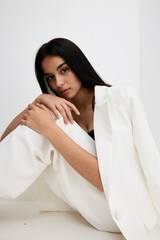 Young woman in white suit sitting on floor with relaxed expression, long dark hair, minimalistic...