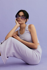 young woman wearing sunglasses and casual sleeveless outfit sitting against purple background with...