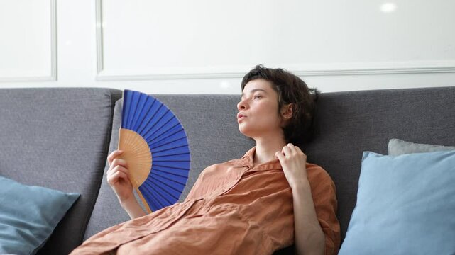 Weary tired woman struggling with sweltering weather using fan to cool down in airless room in summer. Heat exhaustion, physical fatigue, lack of ventilation, heatwave issues, urban living hardship