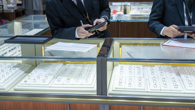 Two jewelry store clerks in formal uniforms inspecting ring displays, using a tablet and clipboard to record inventory inside a brightly lit showcase counter.