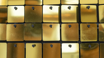 Close-up of a shiny gold sequin wall background with square metallic tiles arranged in a grid pattern, reflecting light and creating a glamorous texture.