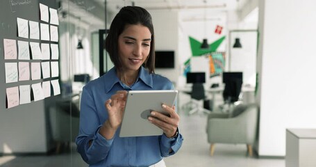 Busy Latin project manager woman typing on tablet at Kanban scrum board with sticky notes, using application for business strategy development, effective management, turning look away, smiling - Powered by Adobe