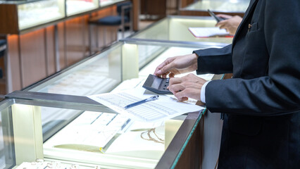 Close-up of a jewelry store staff using a calculator over inventory sheets at a glass display...