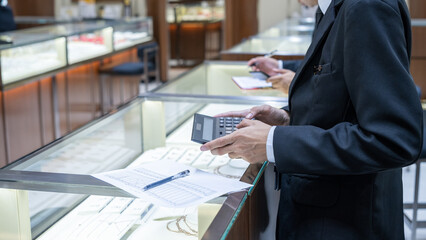 Jewelry store staff in formal attire calculating data with a large calculator and paper sheet near a display case, showing business operations and financial analysis in retail.