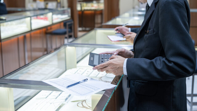 Jewelry store staff in formal attire calculating data with a large calculator and paper sheet near a display case, showing business operations and financial analysis in retail. - Powered by Adobe