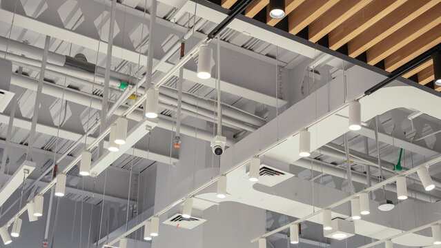 Modern commercial ceiling with industrial-style exposed pipes, hanging LED lights, CCTV camera, air ducts, and fire sprinkler system.