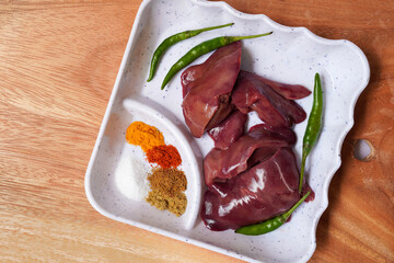 Raw chicken liver on a white plate over wooden background. Fresh ingredients ready for cooking, green chilies, red pepper, turmeric powder. Selective focus and copy space.