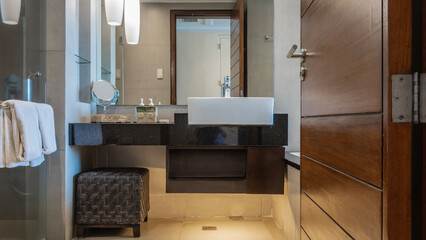 Bathroom. There is a white ceramic washbasin on the marble countertop, dispensers with soap, lotion. A banquette on the floor. The lamps are lit. Towels on the handrail. Reflection in the wall mirror