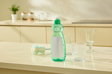 Green dish soap is placed next to glassware on a pale beige counter with gentle light casting soft shadows, giving a calm, fresh vibe. Mockup bottle unlabeled for design. Cleansing