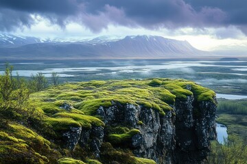 Expansive moss-covered rocky cliff overlooking a misty river delta with distant snow-capped mountains under a moody cloudy sky