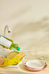 A close-up of dishwashing with soap bottle, yellow gloves, and white dishes under the sink faucet. Clean and organized modern kitchen lifestyle. Mockup bottle unlabeled for design