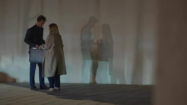 Man and woman exchange briefcase and envelope in urban street deal at night. Illegal transaction or dirty money bribe and fraud. Two people or agents spies meeting in shady suspicious alley to trade