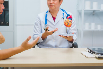 Asian male doctor specializing in cardiology explains heart disease to a patient using a heart model. He provides expert advice, discusses symptoms, risk factors, and treatment in a modern clinic.