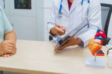Asian male doctor specializing in cardiology explains heart disease to a patient using a heart...