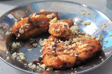 A close-up of golden-brown banana fritters (pisang goreng) drizzled with chocolate sauce and topped with grated cheese, served on a round metal plate. Represents street food, desserts, fusion flavors
