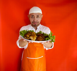 Young Asian Man Chef Using Apron Serving Chicken Roasted On Plate Isolated On Red Background. Studio Shot