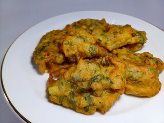 Golden-brown, irregularly shaped fritters with visible green specks, a savory vegetable snack on a white plate with a gold rim. Represents Indonesian cuisine, street food, savory snacks, fried food