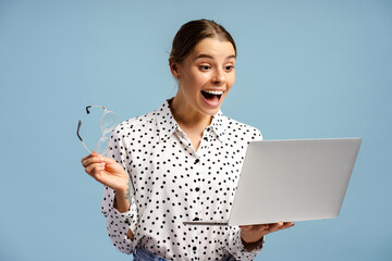 Surprised businesswoman holding eyeglasses and looking at laptop with excitement on blue background