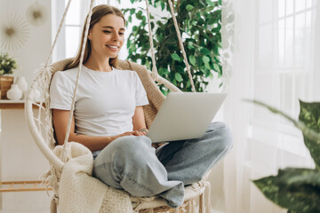 Young smiling woman, freelancer working from home sitting in hanging chair using laptop
