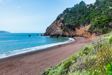 Kirby Cove Beach in California with red sand, blue ocean waves, and dramatic cliffs covered in pine...