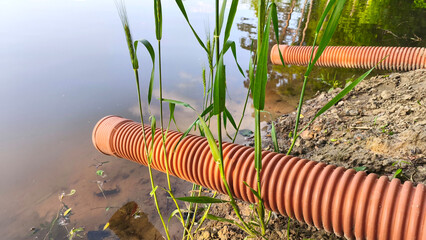 Corrugated drainage pipes lead down into a calm pond from a sandy, eroded bank surrounded by sparse...