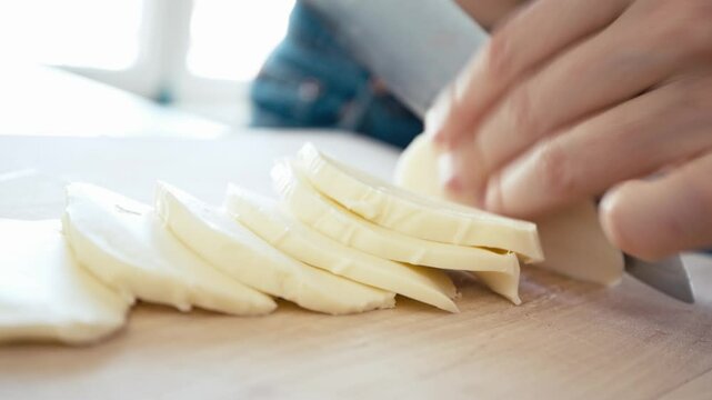 Fresh cheese is being sliced on a wooden board in a sunlit kitchen. The activity showcases skillful hands expertly preparing ingredients for a delicious dish.